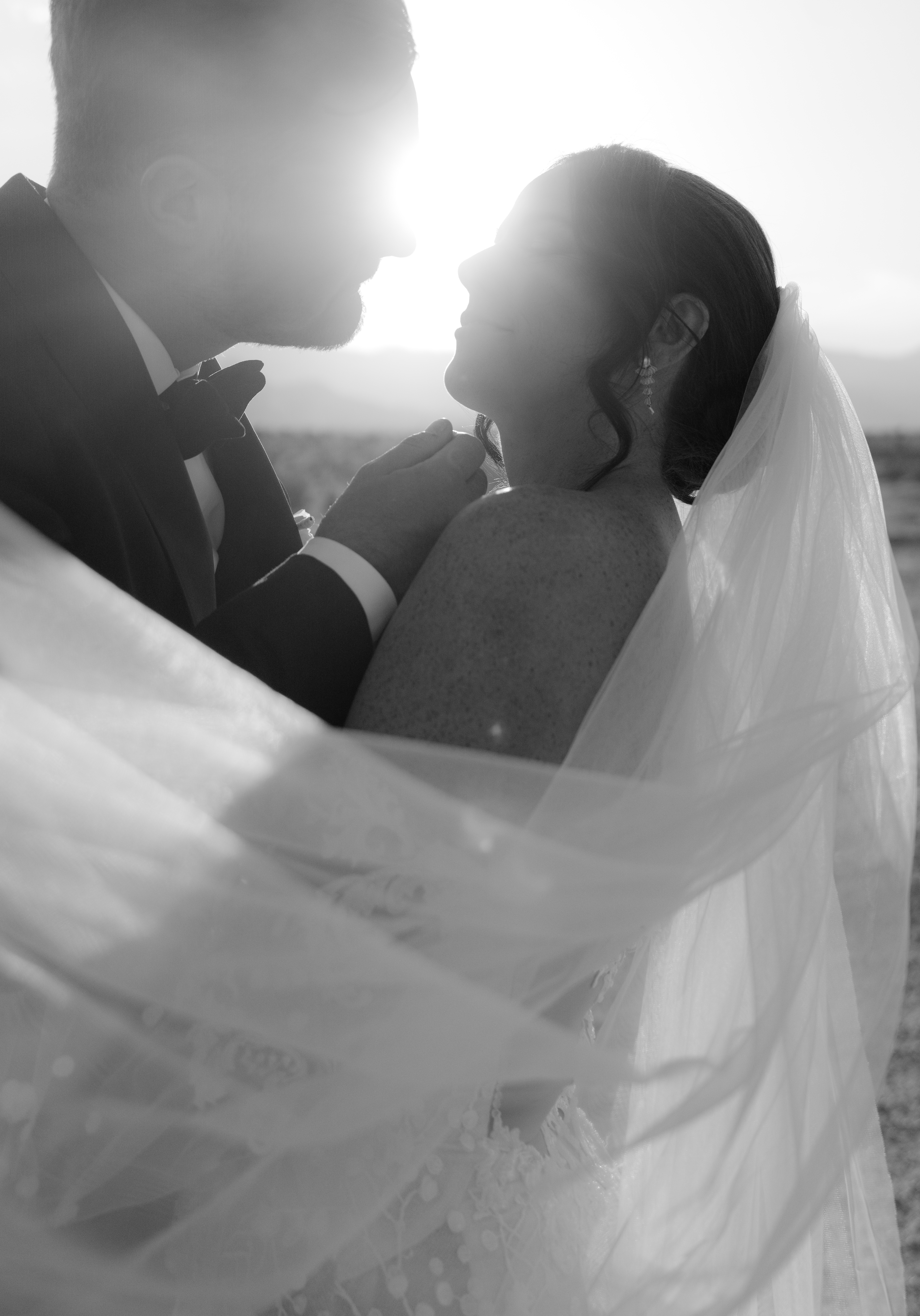 A newlywed couple looks at one another while the sun sets in the background and the brides veil blows in the wind